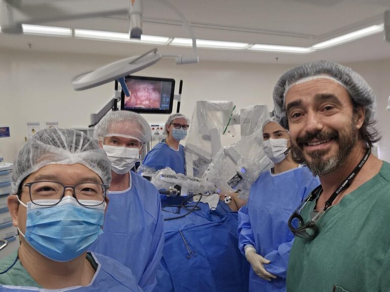 Vale do Paraíba Realiza a Primeira Cirurgia Torácica Robótica da Região 1 Equipe de cirurgiões em uma sala de cirurgia com um monitor ao fundo exibindo parte do procedimento. Estão todos de máscara com excessão de quem tira foto.